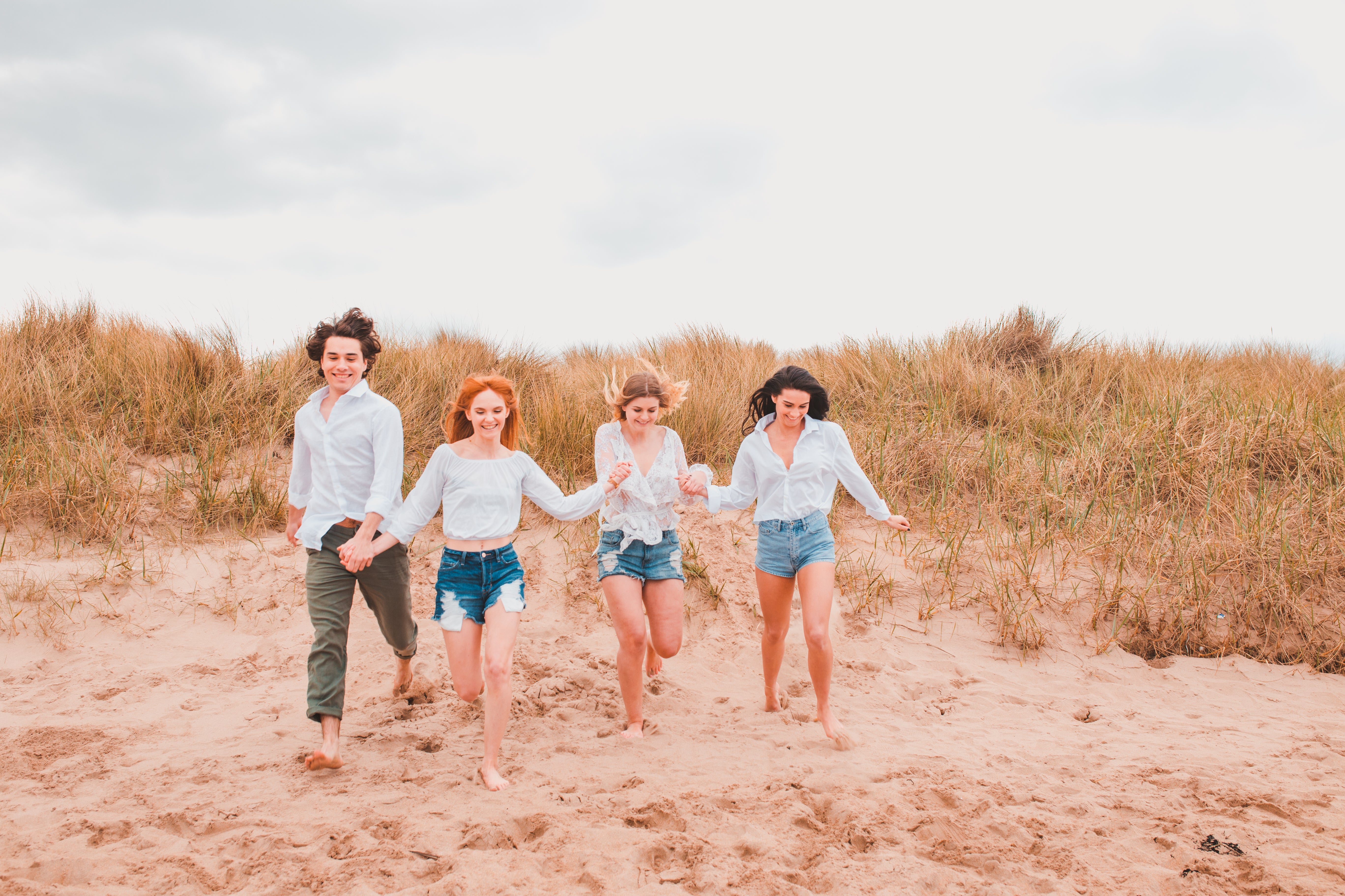 4 models, 3 girls and a boy, are running down a small sand dune towards the camera with their hair flowing in the wind. They are all looking towards the ground and are smiling. There is sand and dune grass in the background. 