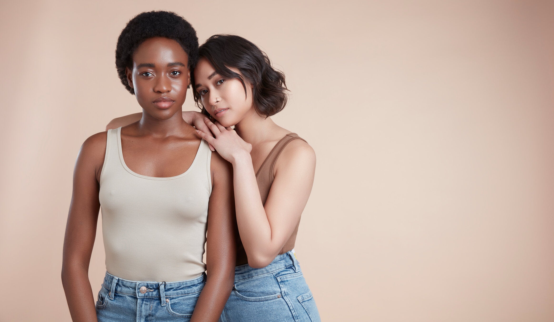 Two female models are looking at the camera with a neutral expression. The model on the left has short black afro hair and the model on the right has a wavy bob. Both models are wearing neutral tops and are standing in front of a neutral backdrop.