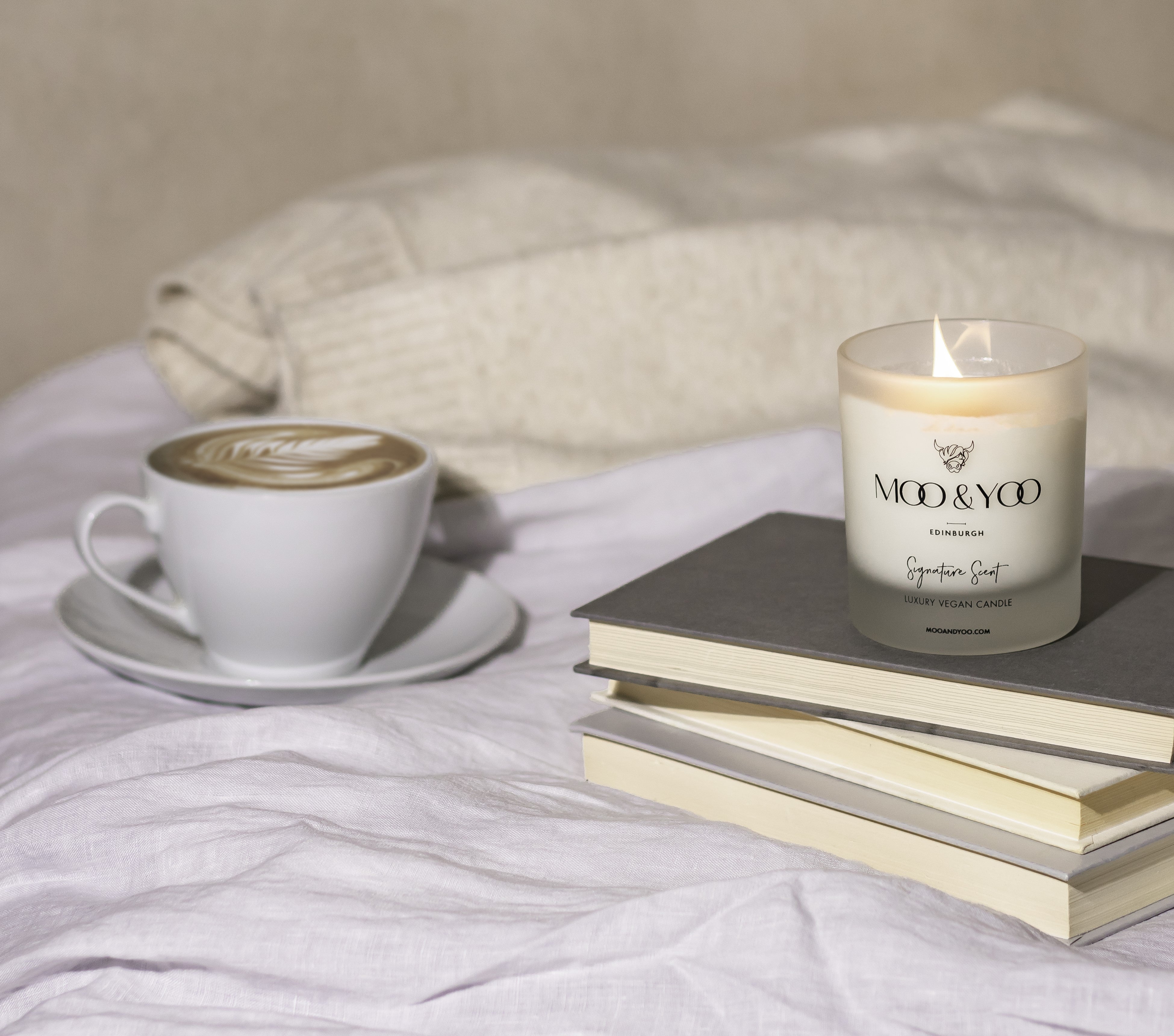 There is a coffee and a Moo & Yoo candle on top of a bed. The candle is on the right hand side of the screen and is balancing on top of three books. The bedding is white and there is a cream coloured throw in the background.