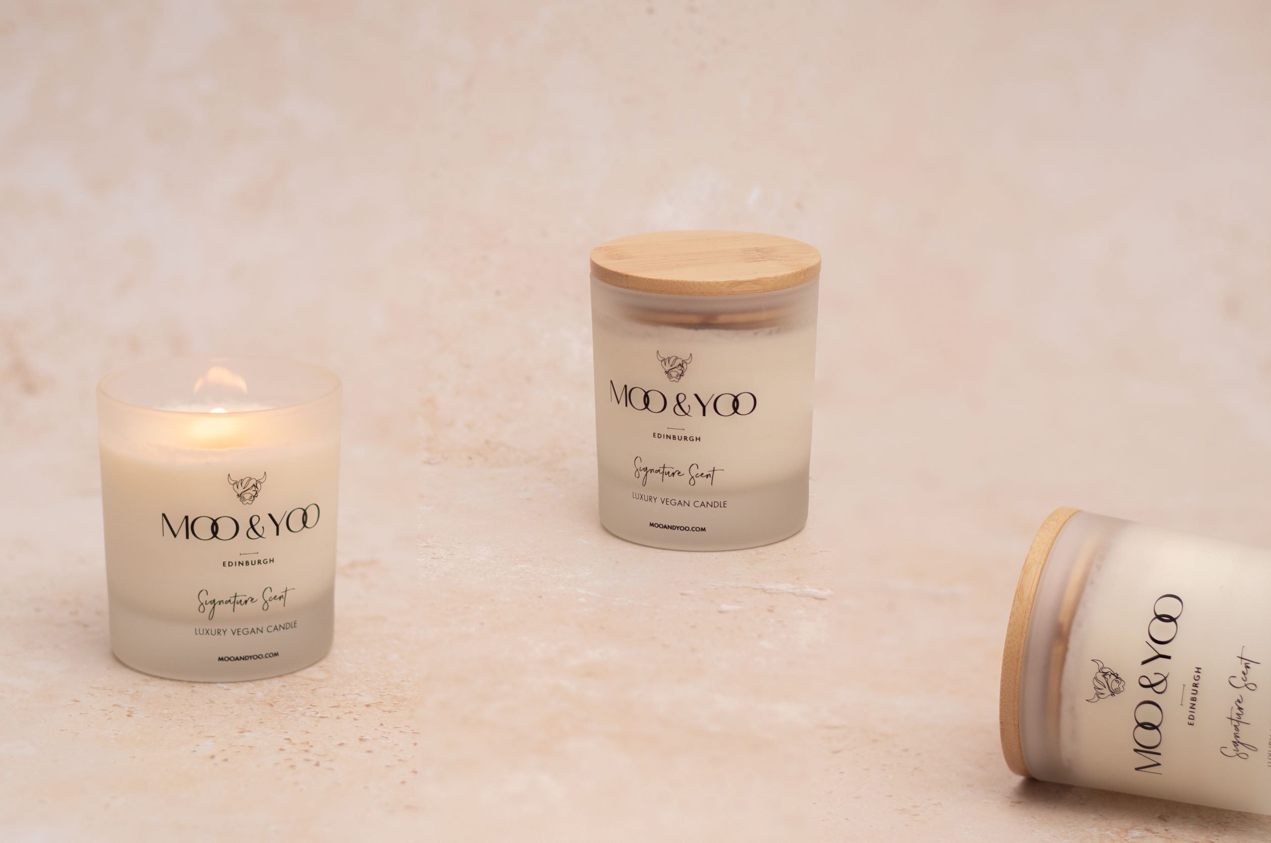 3 Moo & Yoo signature scent candles are places on a speckled nude backdrop. One candle is lit, one candle is stood uprights and the 3rd candle is lying flat.