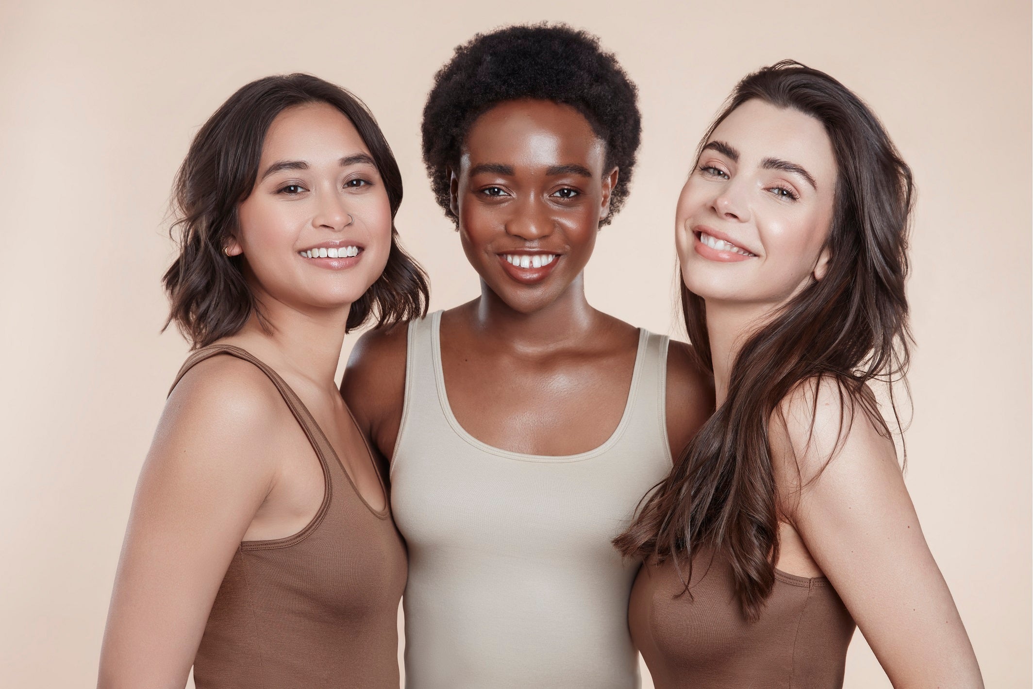 This is a studio image of three female model who are all looking and smiling at the camera. They all have brown hair each varying in texture and length.