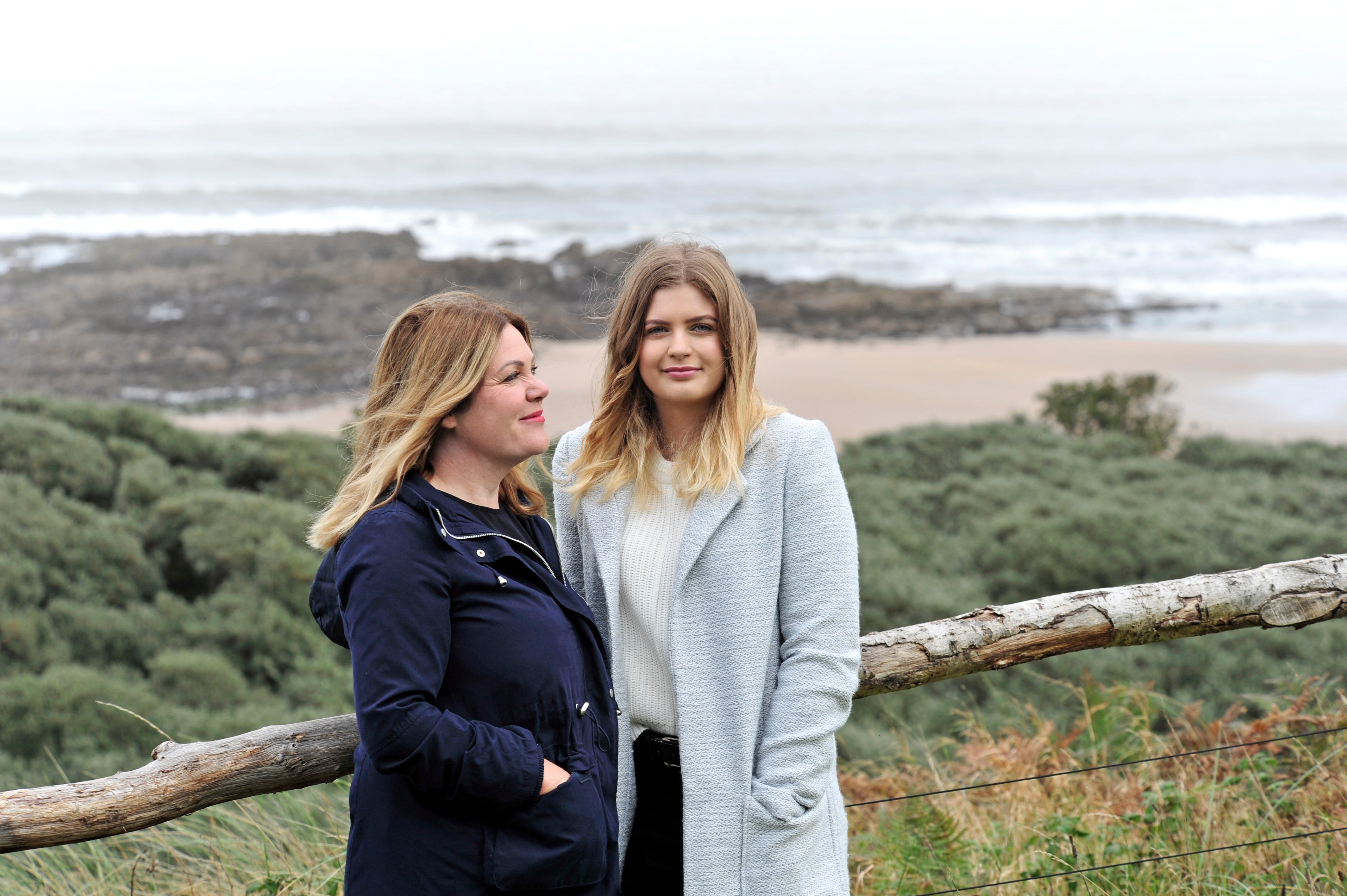 Two women are standing on a hill in front of a beach. The woman on the left has blonde wavy hair and is looking and smiling at the woman on the right. The woman on the right is looking and smiling at the camera. She has blonde wavy hair.