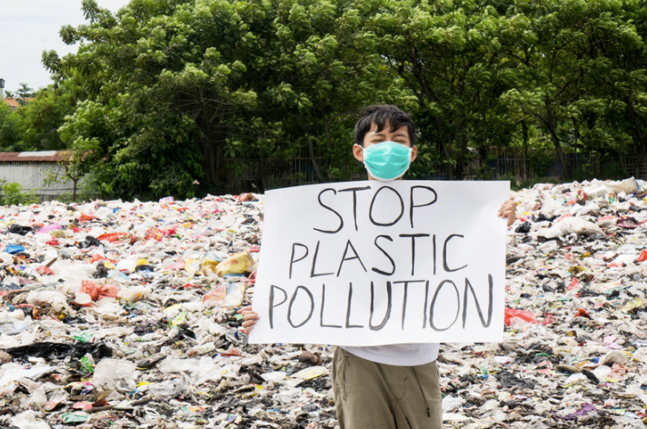 A person is standing in a large pile of plastic waste and is holding a hand written sign that reads 'stop plastic pollution'. The background is plastic waste with some trees in the distance.