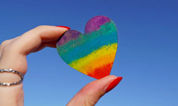 This image shows a hand holding a love heart up to the sky. The love heart is painted with a rainbow on it to represent pride month.
