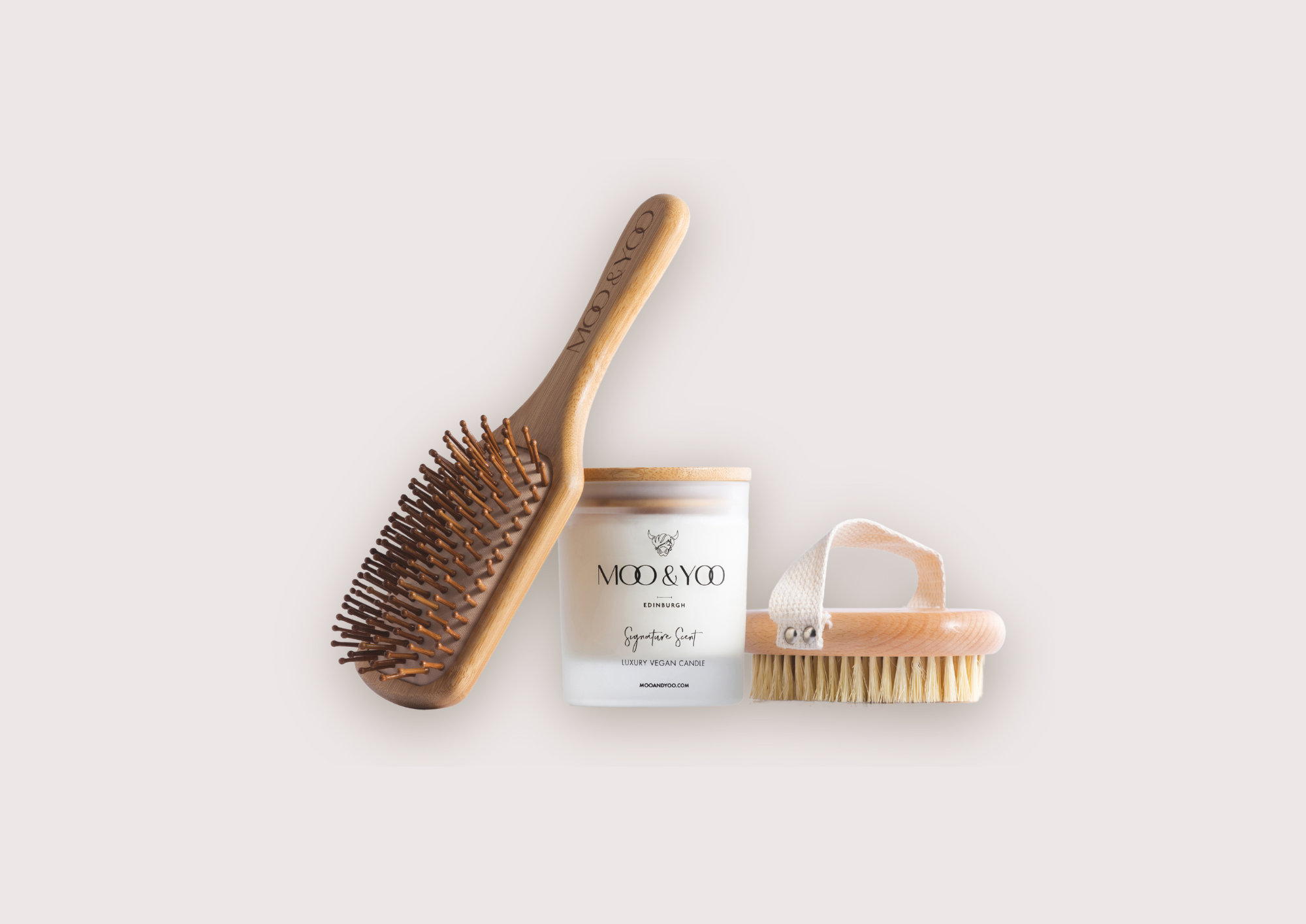 A bamboo paddle hair brush, a Moo & Yoo signature scent candle and a dry body brush and all leaning against each other with a plain background behind them.