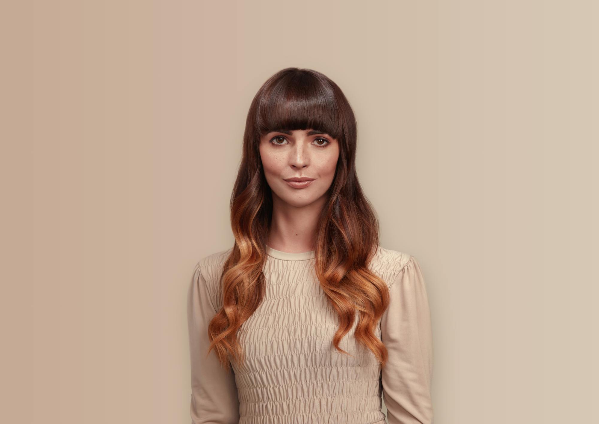 A female model with brown/auburn long flowing hair is standing against a neutral background looking at the camera. She has neutral expression on her face.