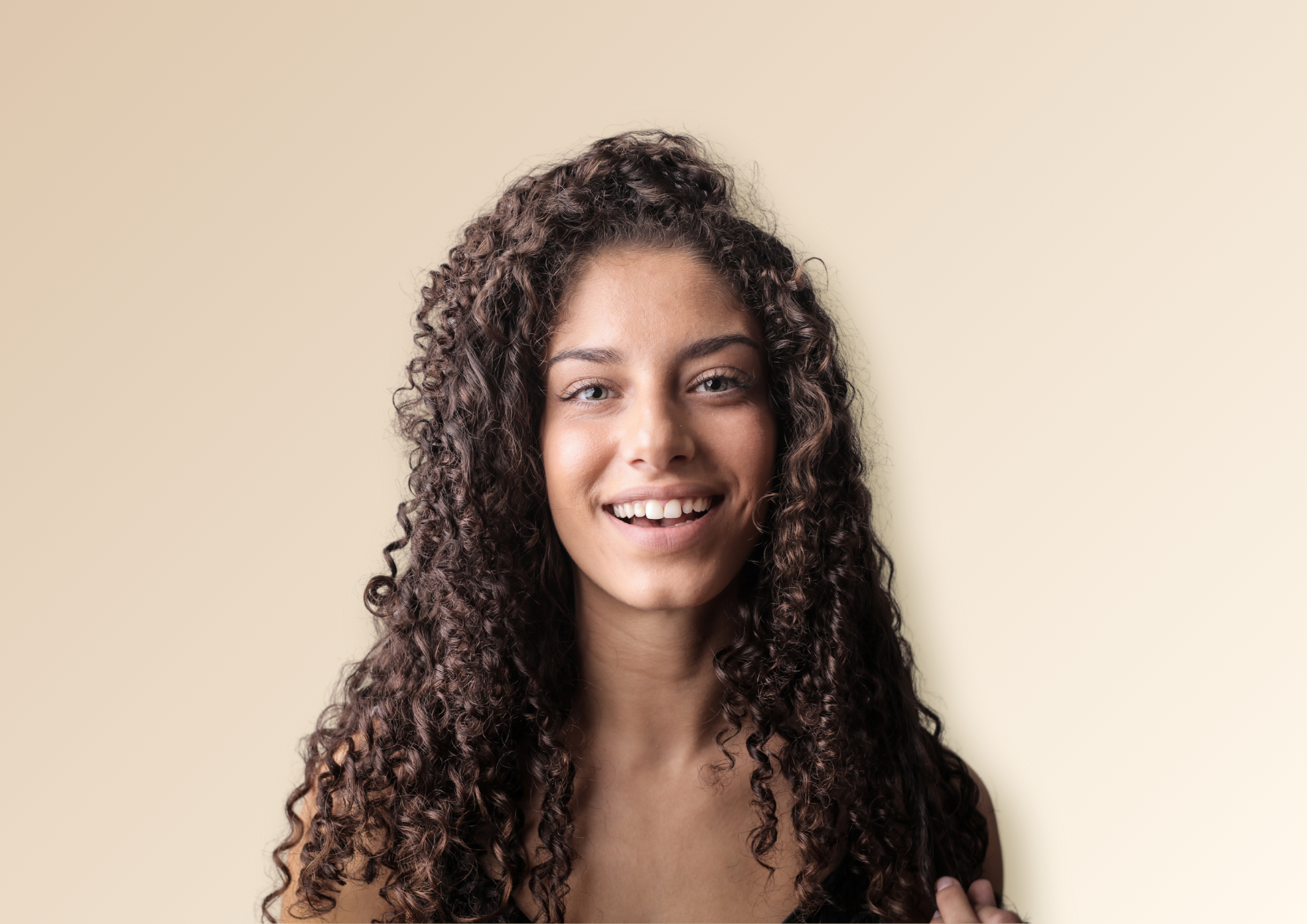 A curl with long, curly, dark brown, type 3B hair is in front of a neutral background and smiling at the camera.