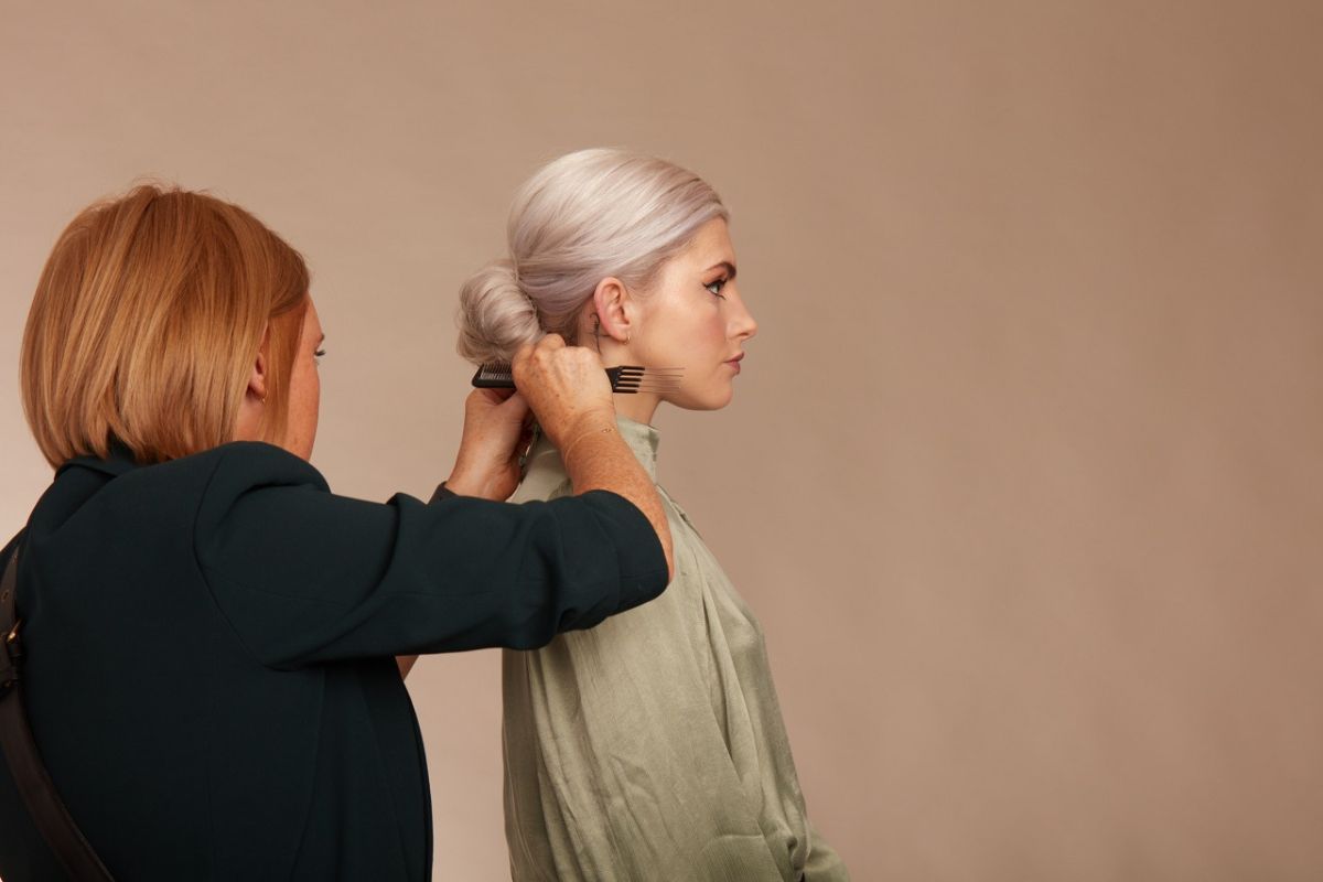 A model with a low blonde bun is standing side on to the camera and there is a hair stylist with a ginger straight bob fixing the models hair.