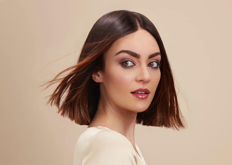 A female model with short, brunette, type 1A hair looking to her left with her hair being blown behind her. She is against a neutral, beige background.