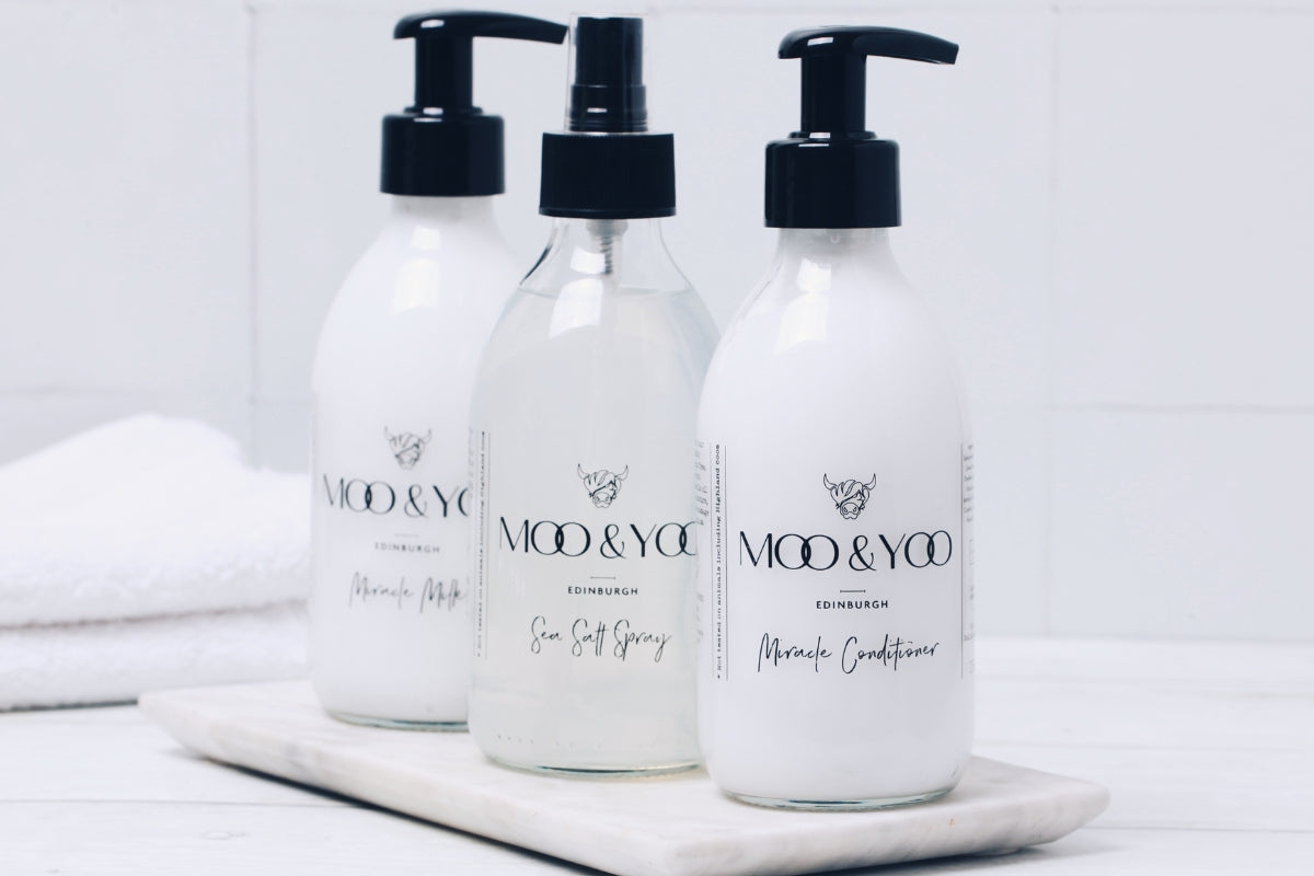 trio of moo glass bottled hair products in a diagonal line on a stone slab in a bathroom setting with white tile background and a white fluffy towel to the left 