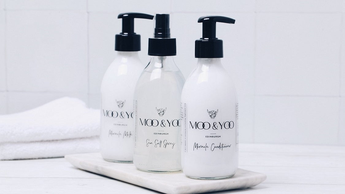 There are 3 Moo & Yoo products (Miracle Conditioner, Miracle Milk and Sea Salt Spray) are sitting on a marble tile which is placed on a white wooden floor. There is a white tiled background with a folded white towel in the background of the products.