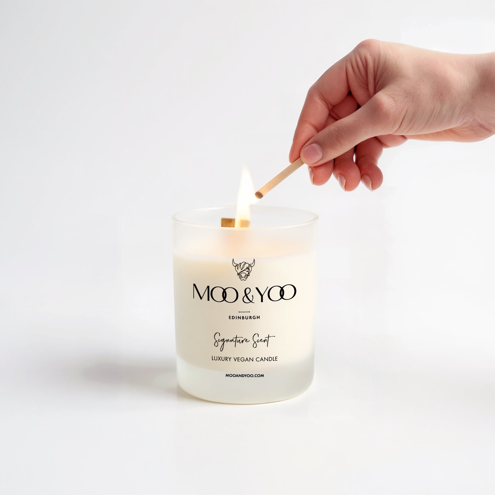 moo & yoo vegan soy wax candle lit with a hand holding a match stick above it against a neutral background 