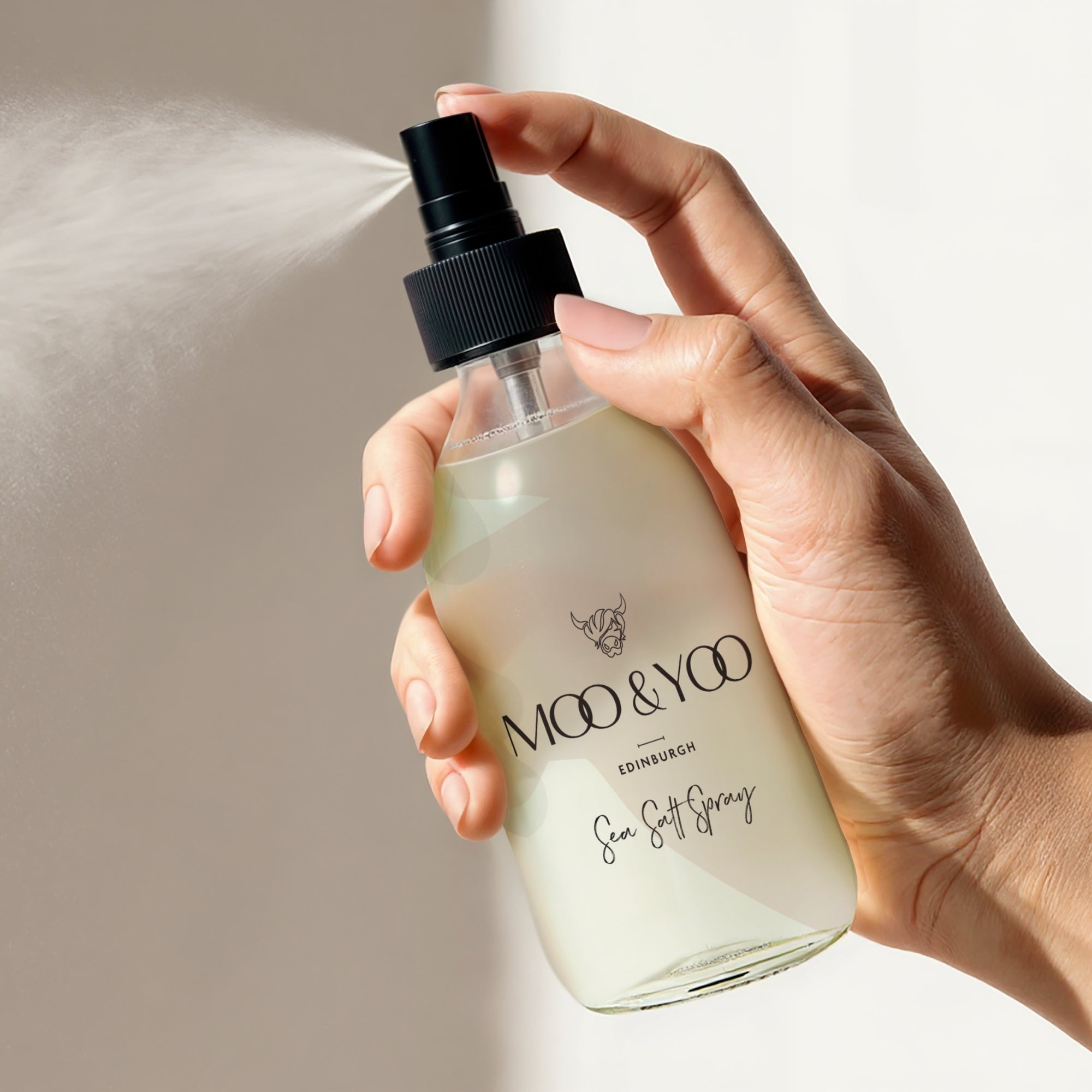 hair care product image of glass moo & yoo salt spray hair styling product in hand being sprayed against a neutral beige background