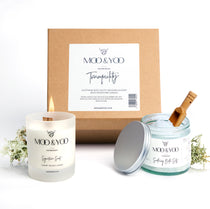 Image of a Kraft natural brown square gift box with label on the front  displaying the gift box name which is Tranquility.  The contents of the box are in front of it, Signature Scent Candle, Soothing Bath Salts and small wooden scoop for the bath salts.