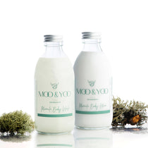 Two glass bottles containing Miracle Body Wash and Miracle Body Lotion sitting side by side on a white bacground with sprigs of moss on either side.