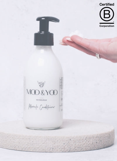 banner with b corp logo on the bottom right, miracle conditioner in a glass bottle sitting on a linen circle platform with a hand outstretching and product on the hand