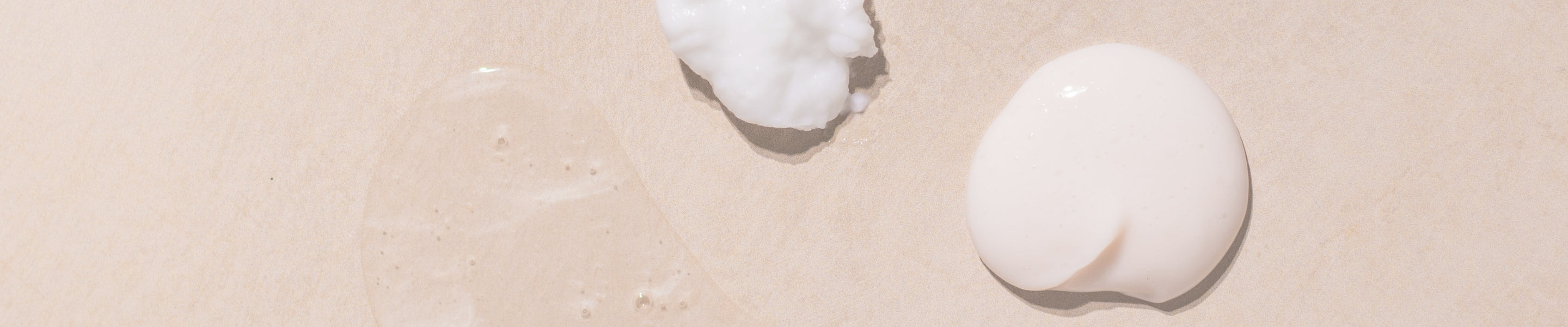 Beige stone surface with three different product textures on the surface. The left is clear liquid with some bubbles which is shampoo, the top middle is thick white cream which is our hair mask and the right is a white smooth lotion texture which is our conditioner 