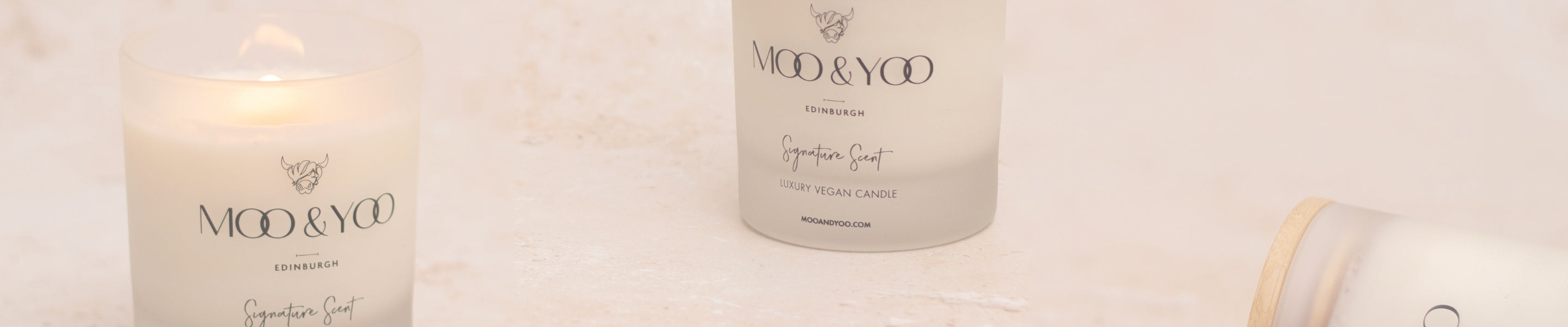 Candles spread pout across image with the candle on the left open and lit, the middle still with the bamboo lid on and the right lying on its side. All vegan candles are branded with 'Moo & You' in glass jars with wooden wicks on a light background