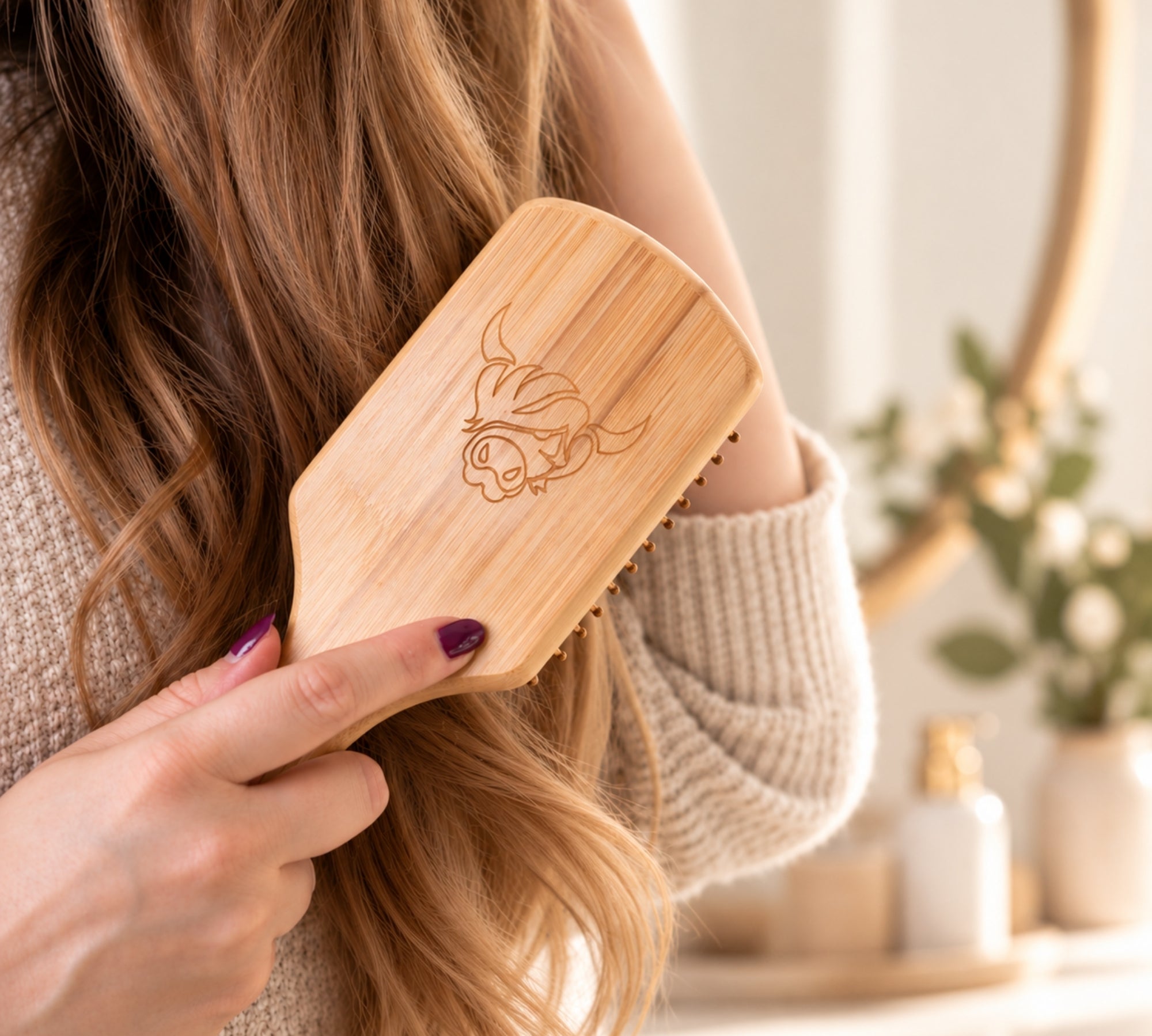A woman with long light brown hair holds a bamboo hairbrush with an engraved highland cow. She is wearing a beige knit sweater in a bright, softly blurred bathroom setting.
