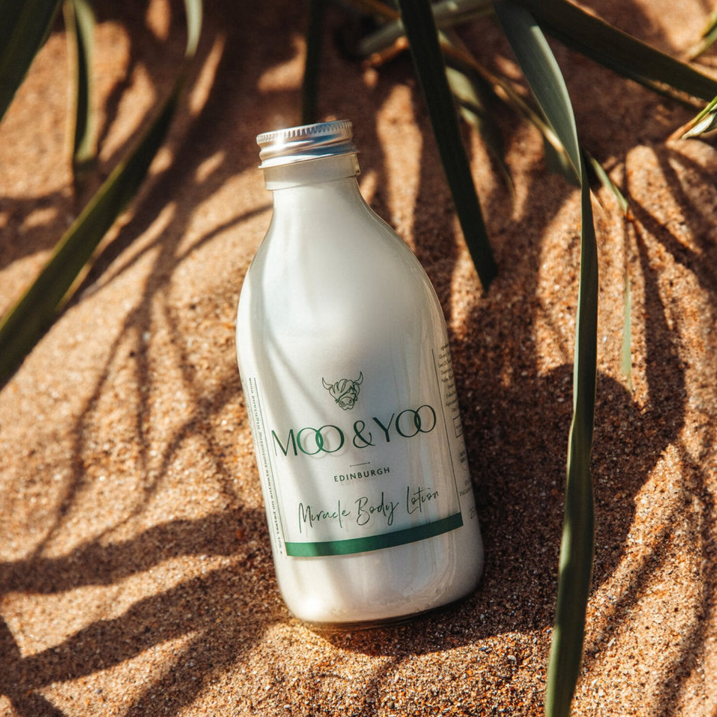 Glass bottle of Moo and Yoo Miracle Body Lotion with aluminium lid on a beach.
