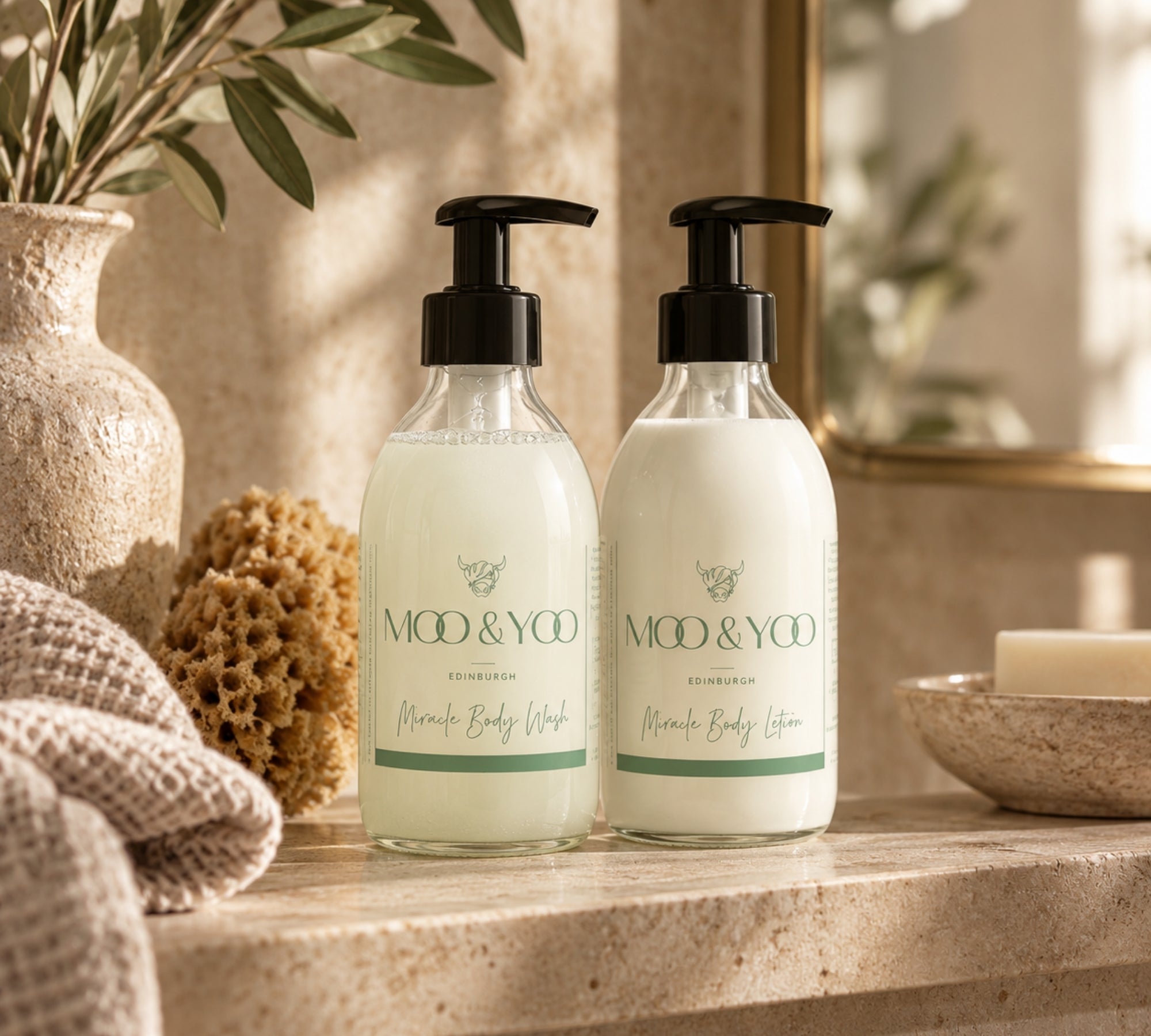 Two pump bottles of MOD & YOO body wash and body lotion standing on a stone surface in a warm bathroom setting, with a vase, burlap sponge, towel, and soap dish nearby.