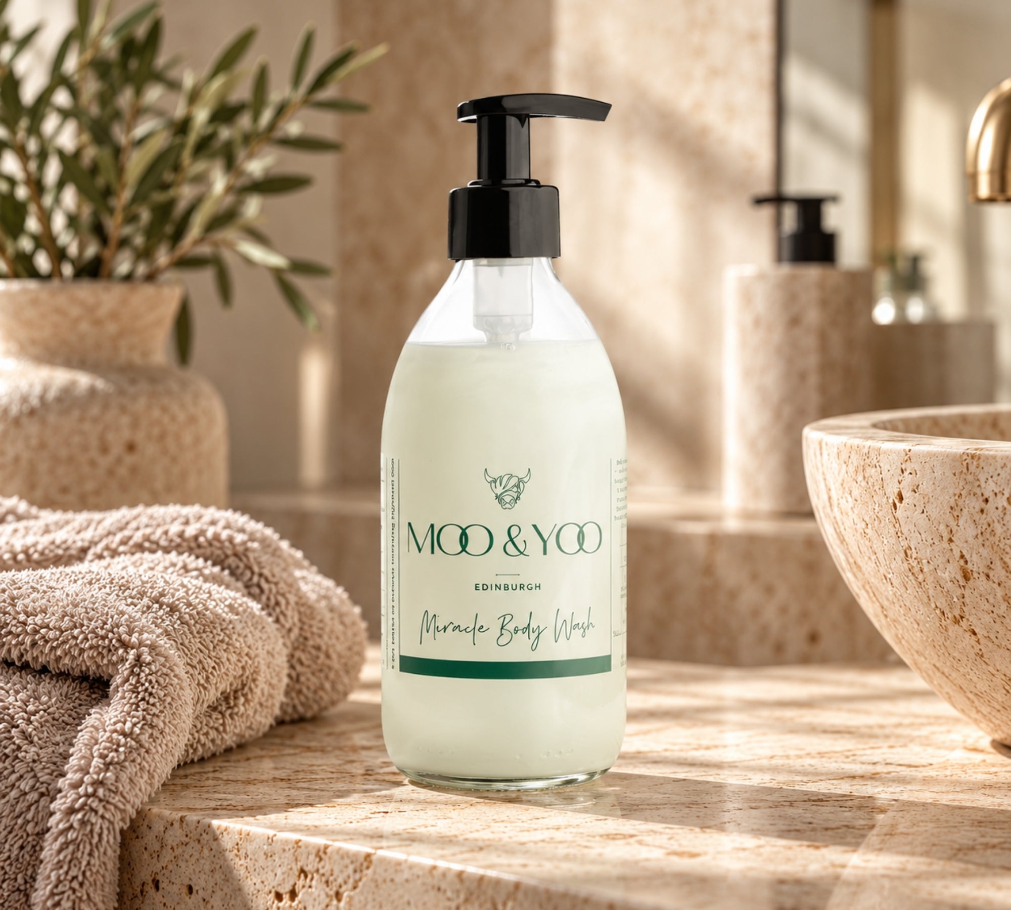 A bottle of MOO & YOO Edinburgh Miracle Body Wash standing on a stone bathroom counter, with a towel, plant, and sink visible in a warm, sunlit setting.