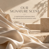 A warm beige promotional graphic with draped cream fabric, stone blocks, and shadow play, plus the text 'OUR SIGNATURE SCENT' and a short descriptive blurb about a Cashmeran blend with floral and botanical notes.