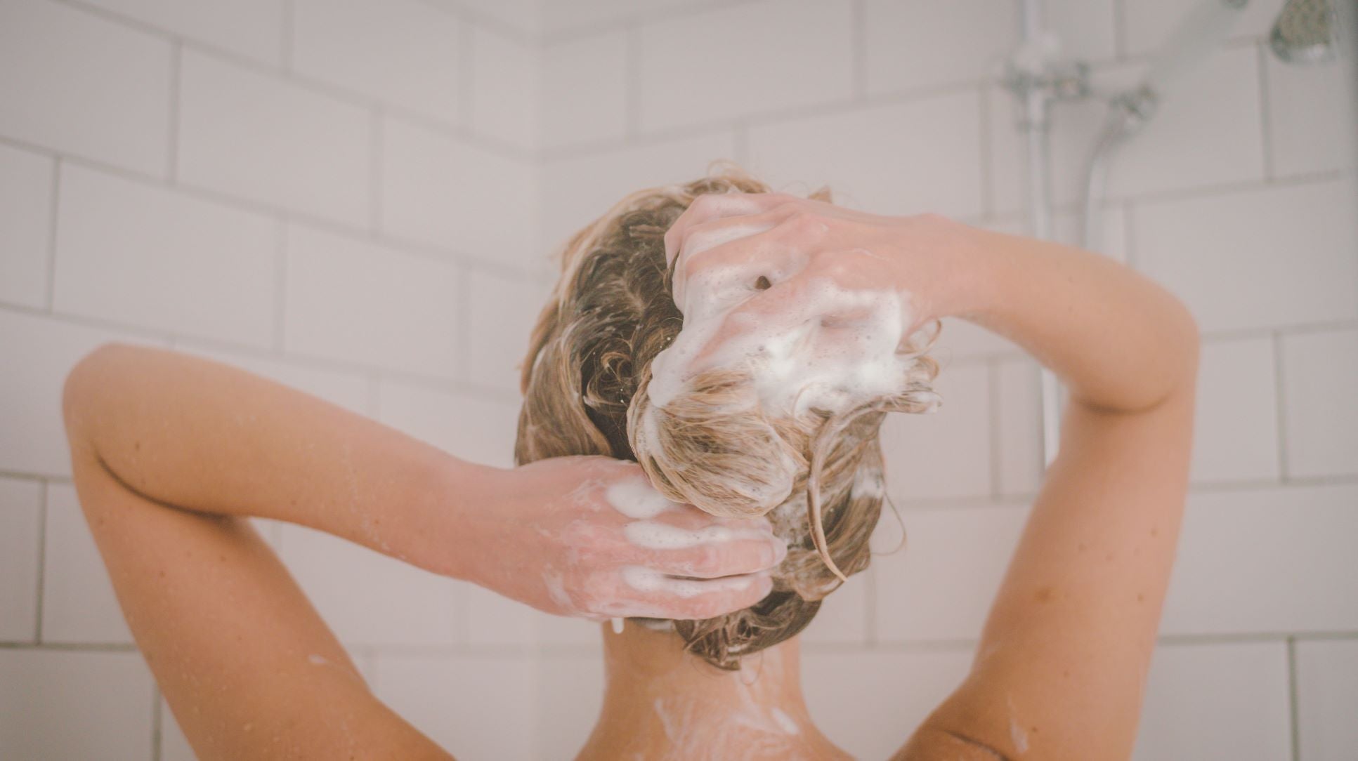 The back of a girls head in the shower, her hair pulled up covered in bubbled from the lather of our miracle shampoo. Her hands are in her hair and she is facing the back shower wall of white subway tiles 