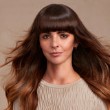 A woman with long, wavy brown hair and straight bangs wearing a beige smocked top, shown against a solid beige background.