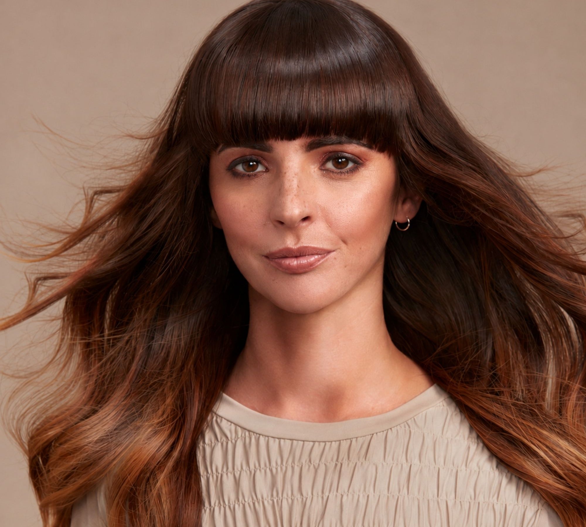 A woman with long, wavy brown hair and straight bangs wearing a beige smocked top, shown against a solid beige background.