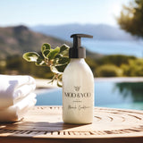 A close up image of a glass bottle of Moo and Yoo Miracle conditioner with a pump. It is placed on a table at a villa overlooking the swimming pool. There is a towel to the side.  In the distance you can see hills and the sea