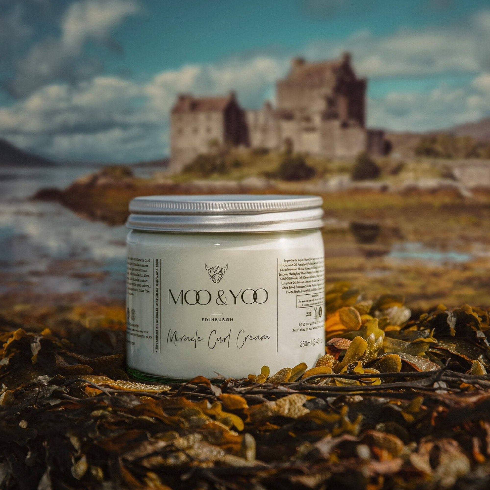 A glass jar of Moo and Yoo Miracle Curl Cream with an aluminium lid. It is sitting on some seaweed on the side of a Scottish Loch with a castle in the background.