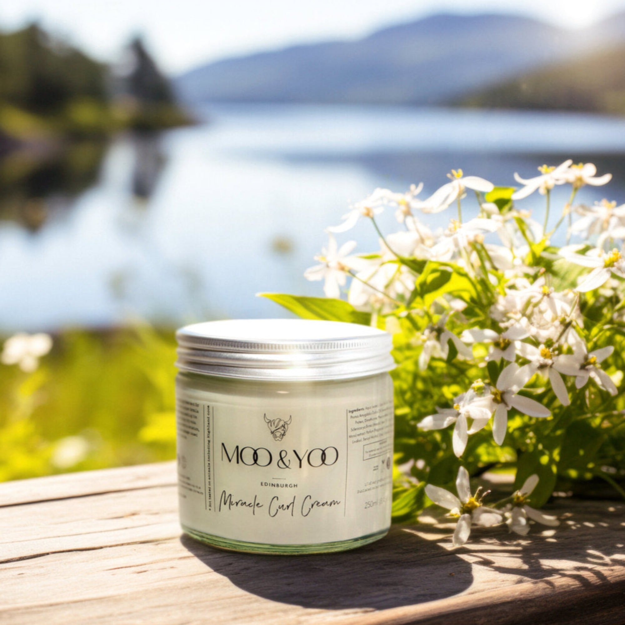 A glass jar of Moo and Yoo Miracle Curl Cream with an aluminium lid placed on a rustic wooden bench overlooking a Scottish Loch and hills. It is a spring day and there are white spring flowers behind it.