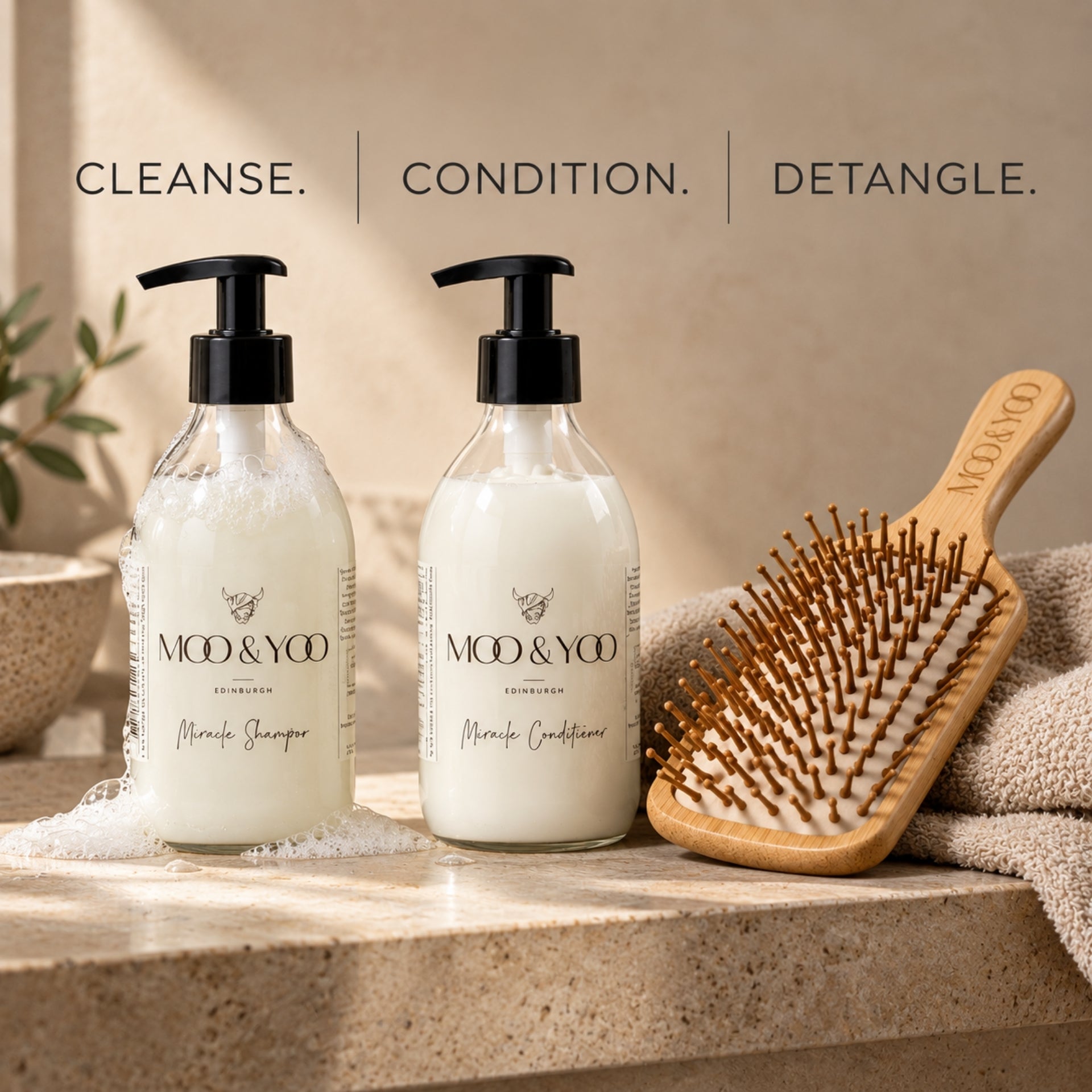 Two clear pump bottles of MOO & YOO haircare products, labeled Mo & Yoo Shampoo and Moo & Yoo Conditioner, displayed on a stone surface next to a wooden bamboo paddle hairbrush and a towel. Text at the top reads 'CLEANSE. CONDITION. DETANGLE.