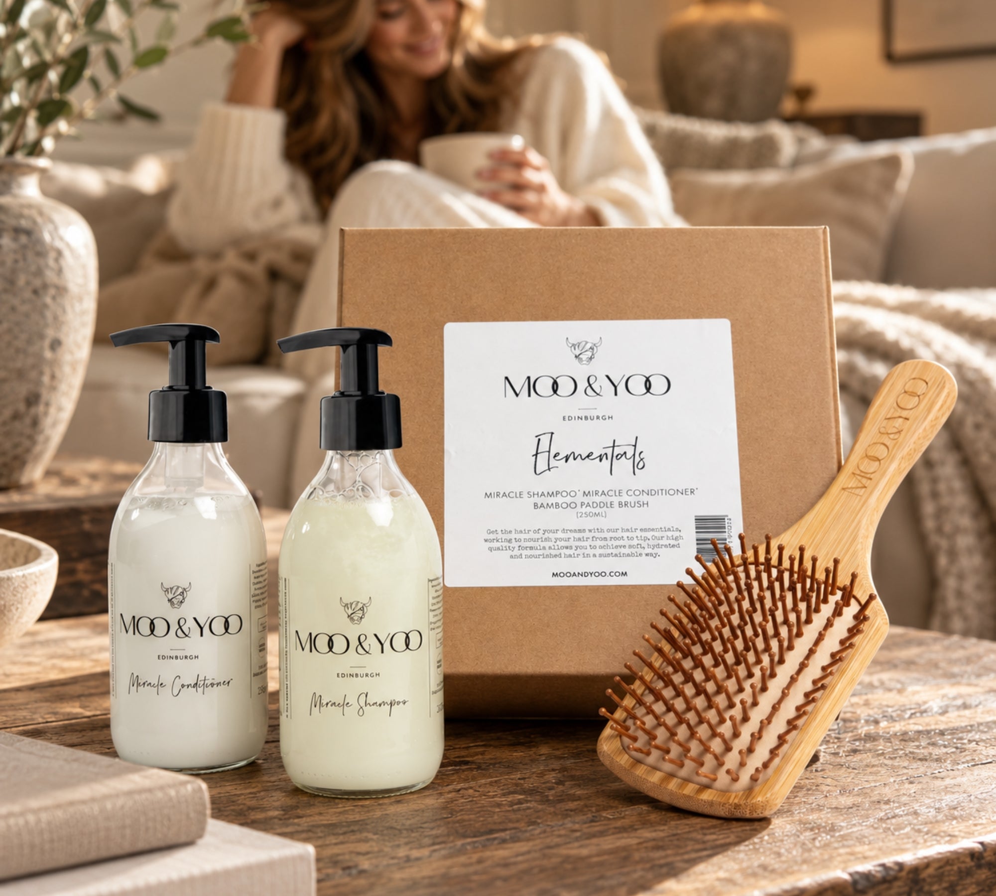 A hair care set from Moo & Yoo. on a rustic wooden table, featuring a kraft gift box, a wooden hairbrush, a  bottle of moo & yoo conditioner with black pump, and a moo & yoo shampoo bottle with a black cap.