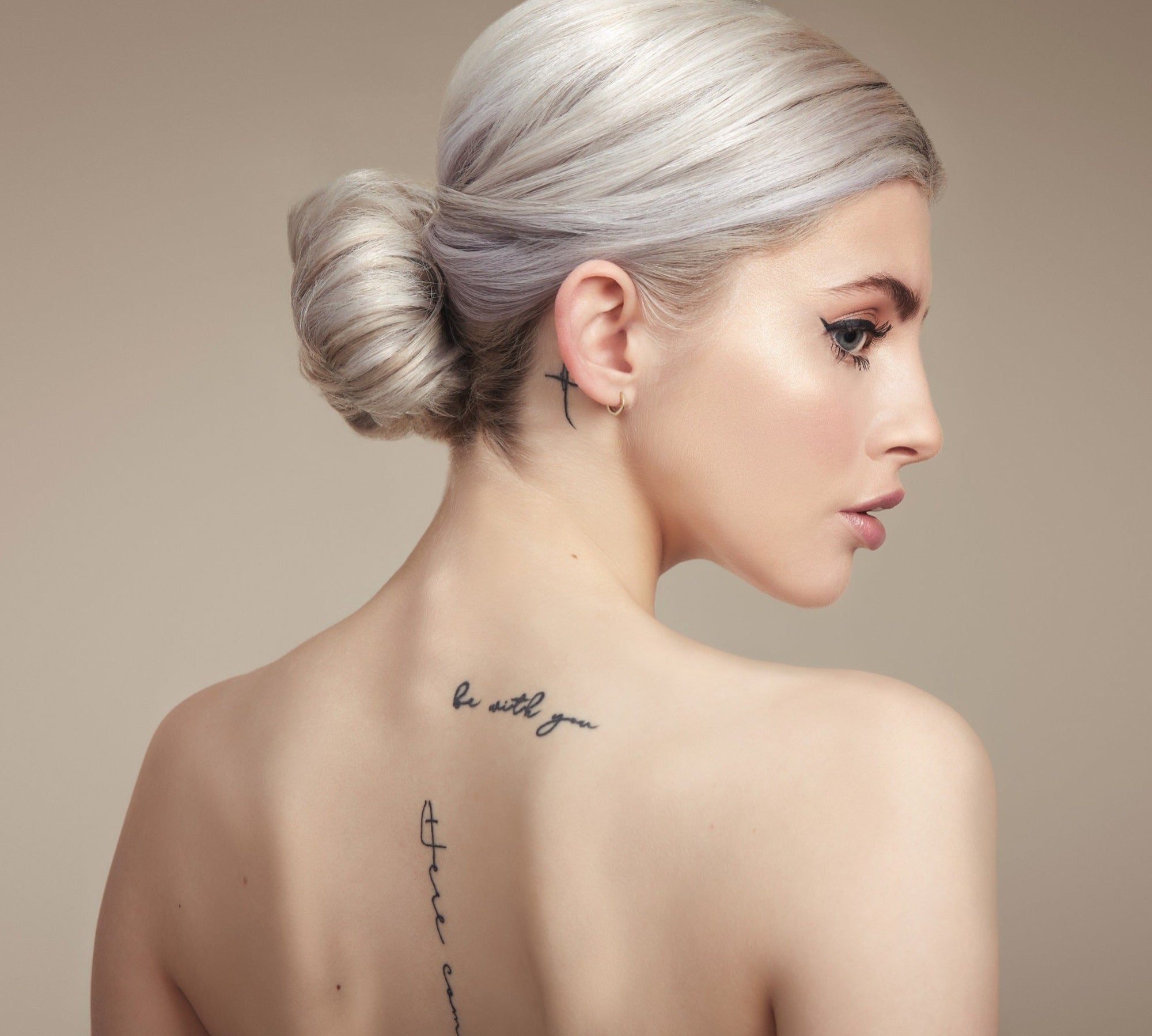 Female model with her back to the camera. Her platinum blonde hair is in a neat low bun, back is bare and she has tattoos down her spine 