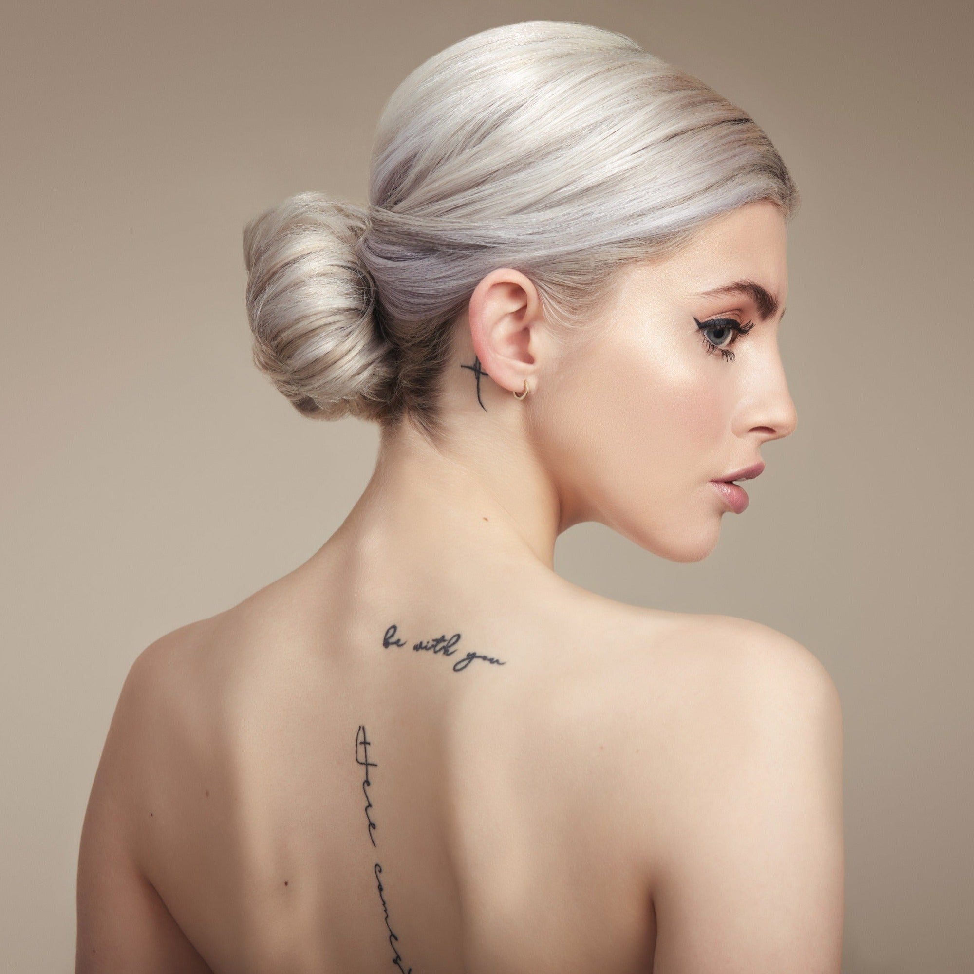 Female model with her back to the camera. Her platinum blonde hair is in a neat low bun, back is bare and she has tattoos down her spine 