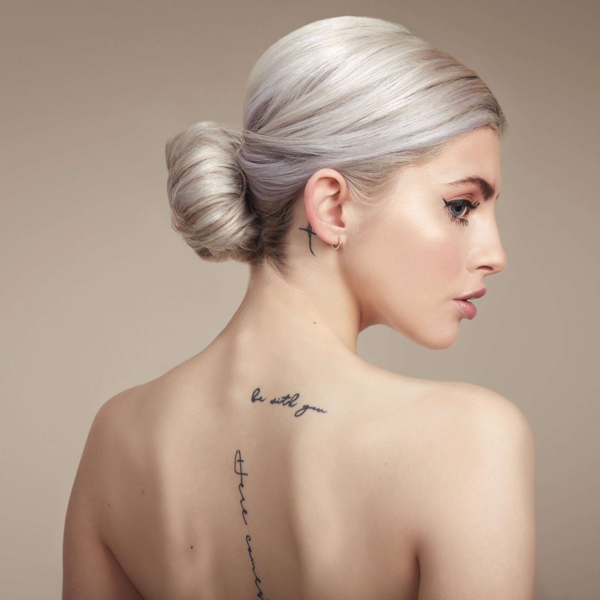 Female model with bleached blond hair in a bun. She has her back to the camera so you can see the bun and she is turning her head to the side so you can see her profile. Her back is bare and she has some tattoos. 