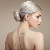 Female model with bleached blond hair in a low bun. She has her back to the camera so you can see the bun and she is turning her head to the side so you can see her profile. She has processed hair so has used Moo and Yoo Miracle Conditioner and Mask. Her back is bare and she has some tattoos. 