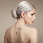 Female model with bleached blond hair in a low bun. She has her back to the camera so you can see the bun and she is turning her head to the side so you can see her profile. She has processed hair so has used Moo and Yoo Miracle Conditioner and Mask. Her back is bare and she has some tattoos. 