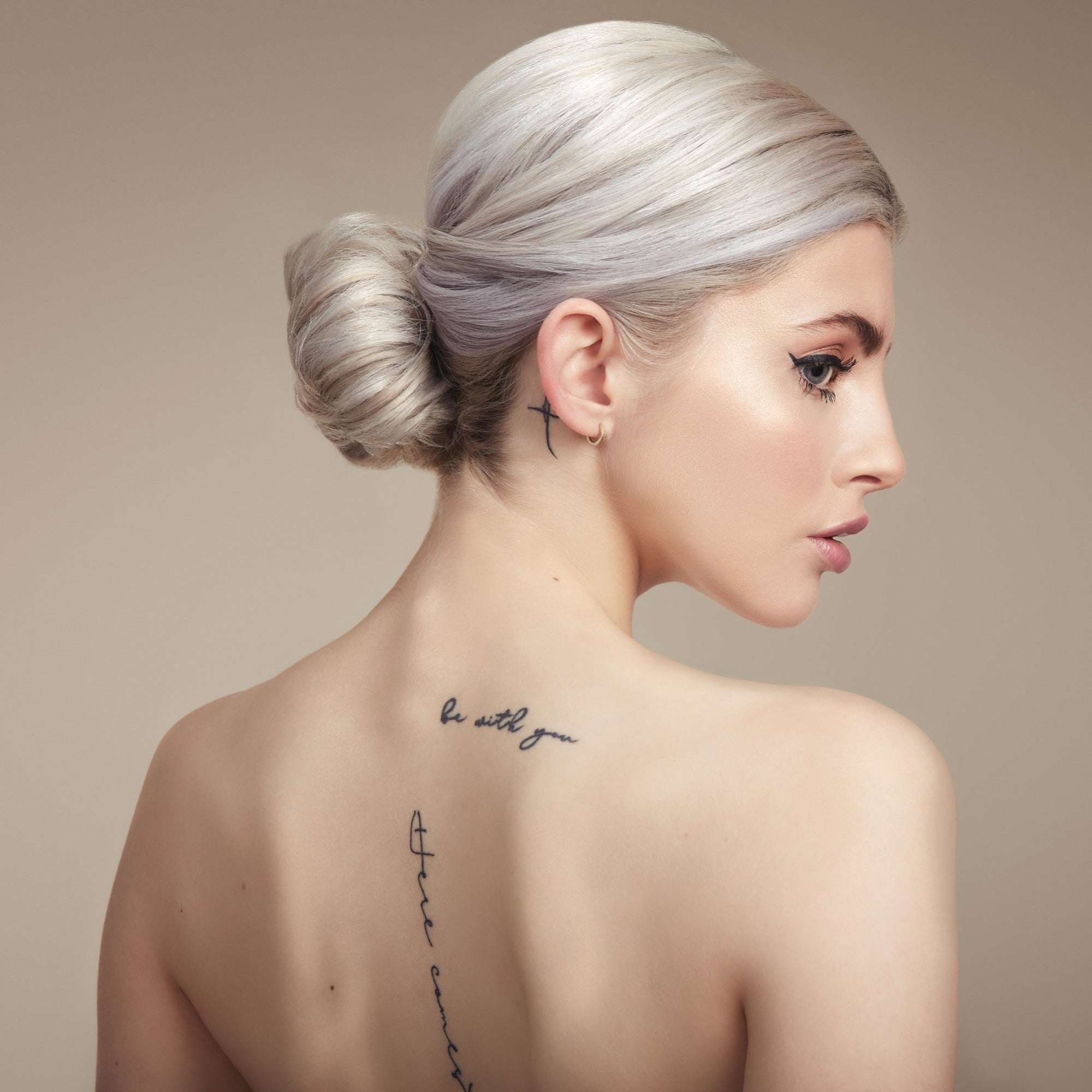 Female model with bleached blond hair in a bun. She has her back to the camera so you can see the bun and she is turning her head to the side so you can see her profile. Her back is bare and she has some tattoos.
