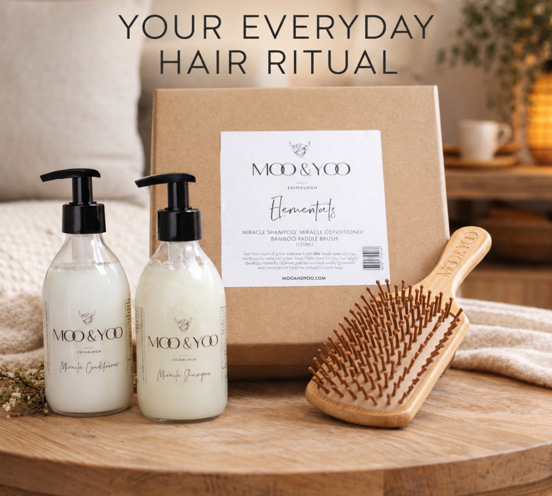 A hair care gift set on a wooden tray, featuring two frosted pump bottles of Moo & Yoo Miracle Shampoo and Miracle Conditioner, a brown kraft box with a white label, and a wooden hairbrush. The background shows a cozy bedroom setting with a bed and a lamp, and the text 'YOUR EVERYDAY HAIR RITUAL' is displayed at the top.