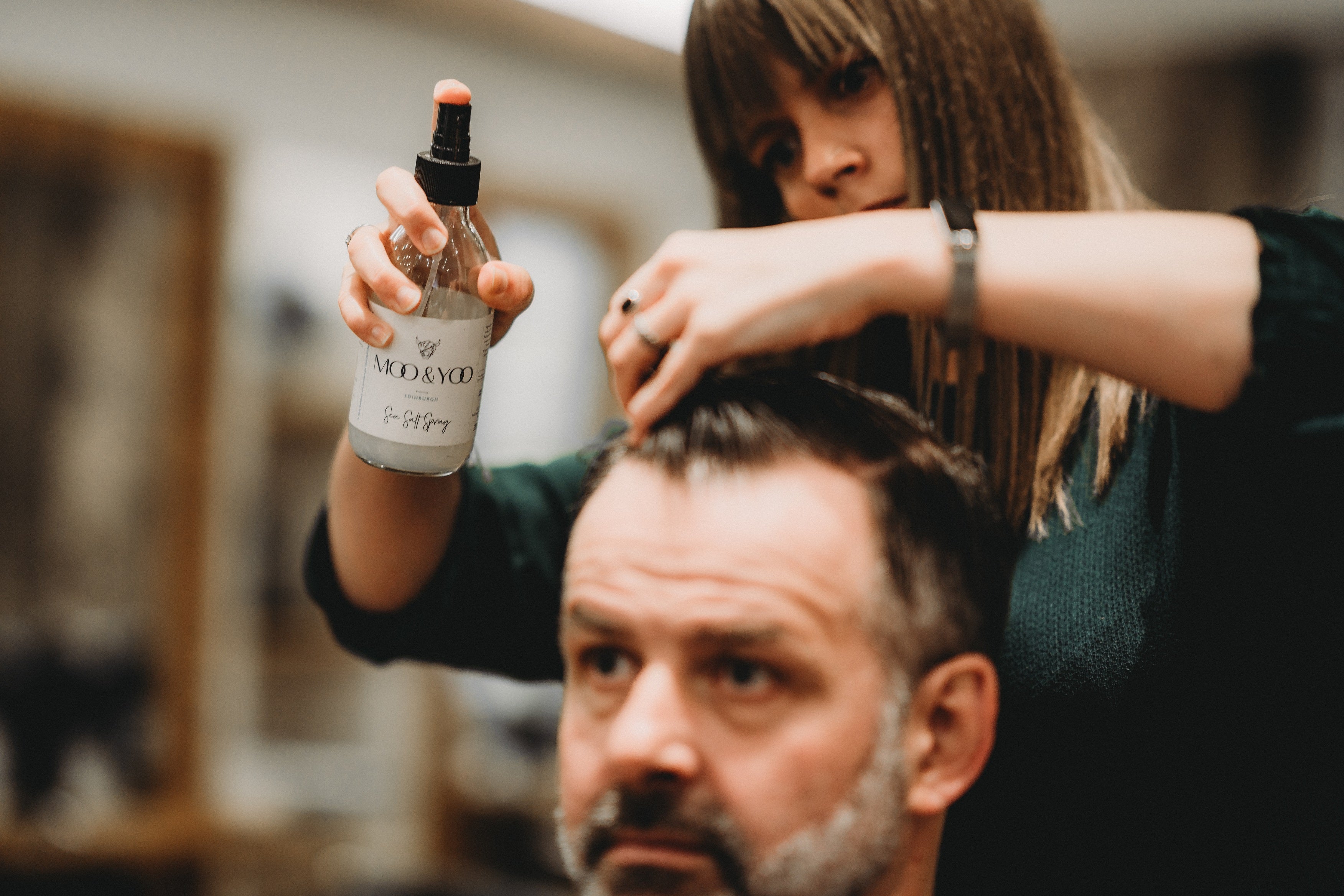 Our sea salt spray being sprayed into a mans hair by a female hair stylist. He has cropped dark hair and she is styling it with her free hand.