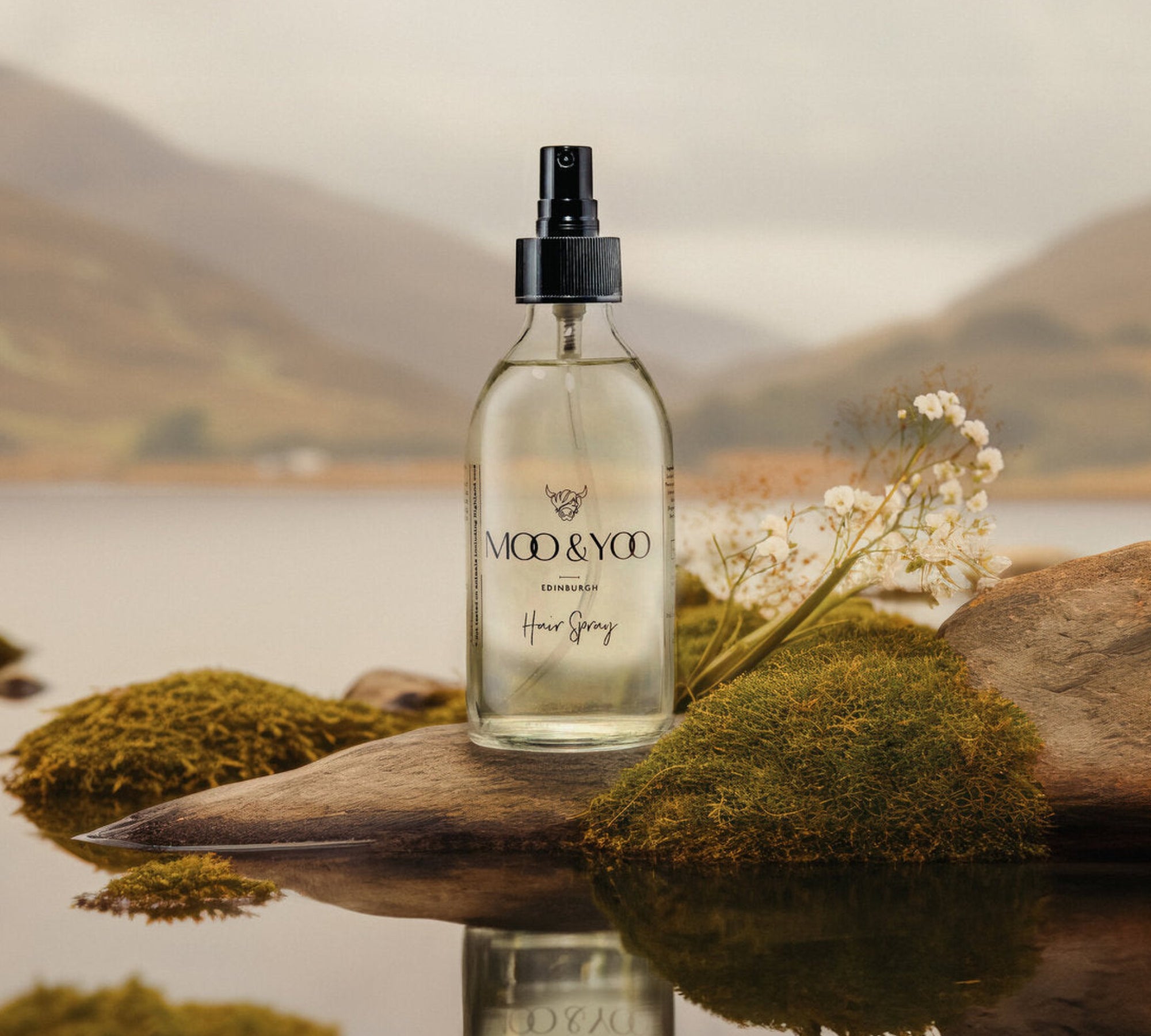 A glass bottle of Moo and Yoo hair spray sitting on a rock with moss at the edge of a Scottish Loch.  It is autumn colours on the loch and hillsides in the distance.