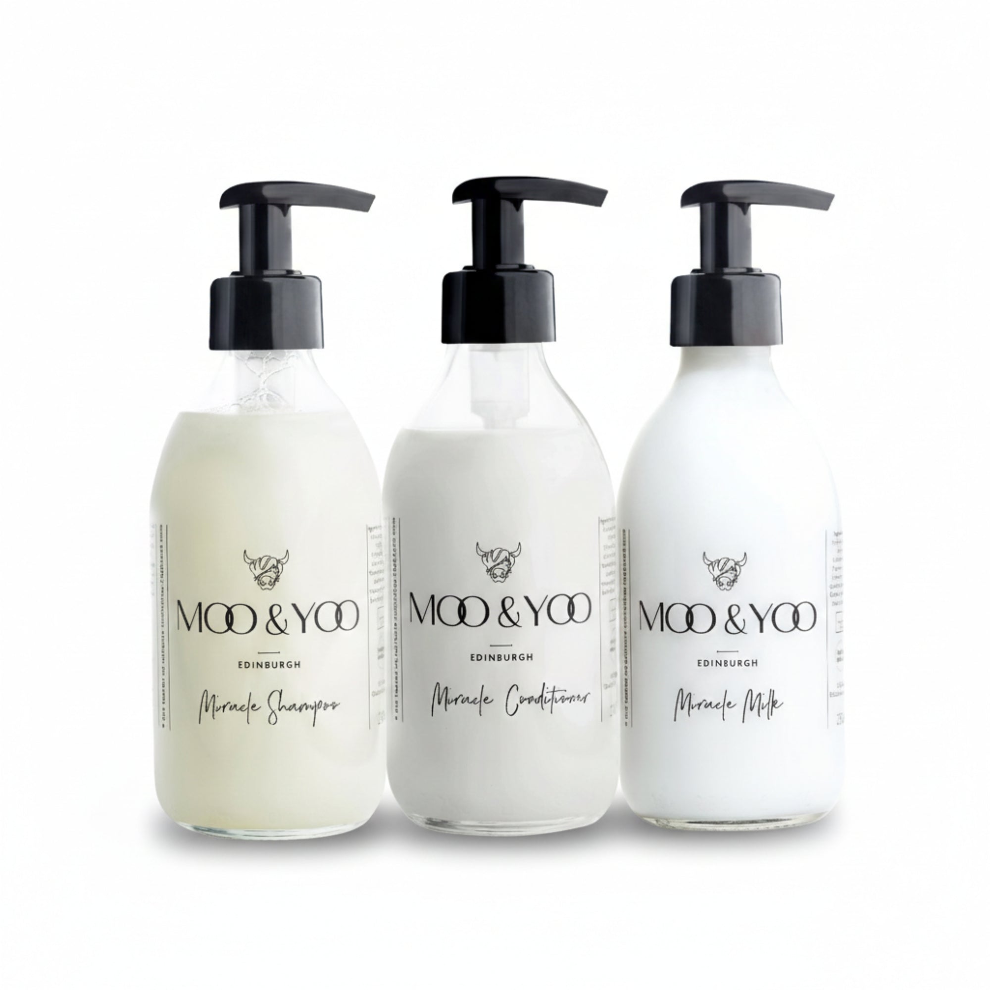 Three glass bottles of Moo and Yoo products sitting side by side. It is a Miracle Shampoo, Conditioner and Miracle Milk leave-in conditioner. They are on a white background