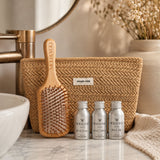 A hair care gift set featuring three silver mini bottles of MOO & YOO hair products, a natural wood hairbrush, and a woven beige tote bag, arranged on a marble bathroom counter next to a sink and dried flowers.