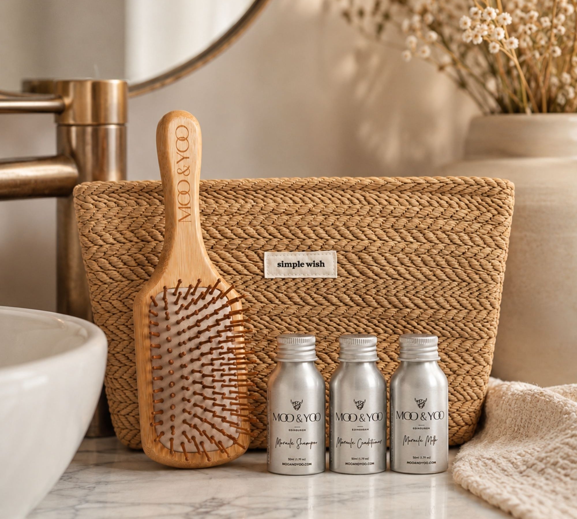 A hair care gift set featuring three silver mini bottles of MOO & YOO hair products, a natural wood hairbrush, and a woven beige tote bag, arranged on a marble bathroom counter next to a sink and dried flowers.