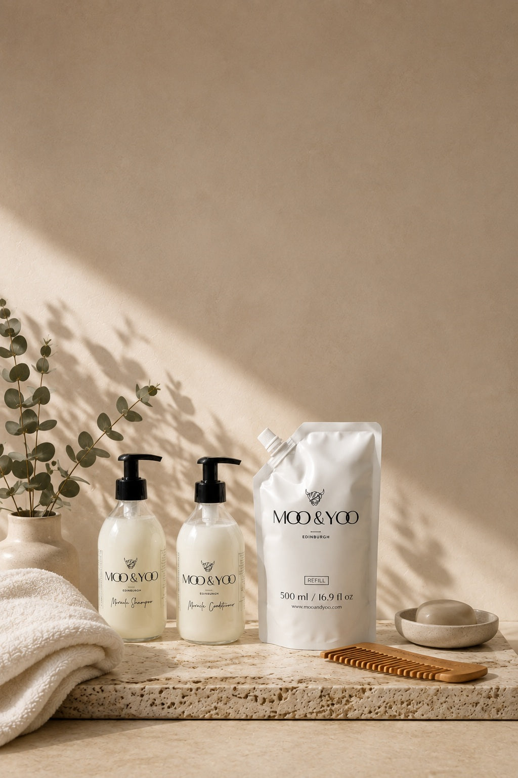 Three MOD & YOO haircare products arranged on a stone tray with a beige background: a large white refill pouch, a white pump bottle, and a smaller white bottle, styled with a folded towel, eucalyptus branch, wooden comb, and soap dish.