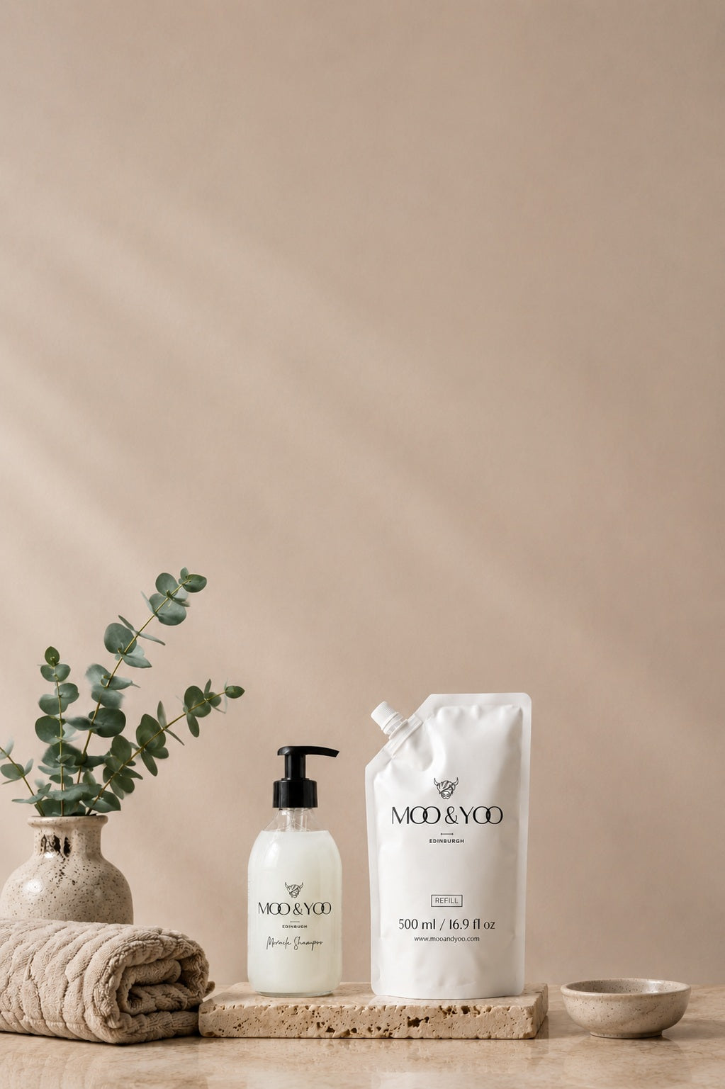 Moo & Yoo Miracle Shampoo and 500ml refill pouch styled on a stone surface with a folded towel, eucalyptus in a ceramic vase and a taupe background, highlighting the brand's luxury sustainable haircare refill system