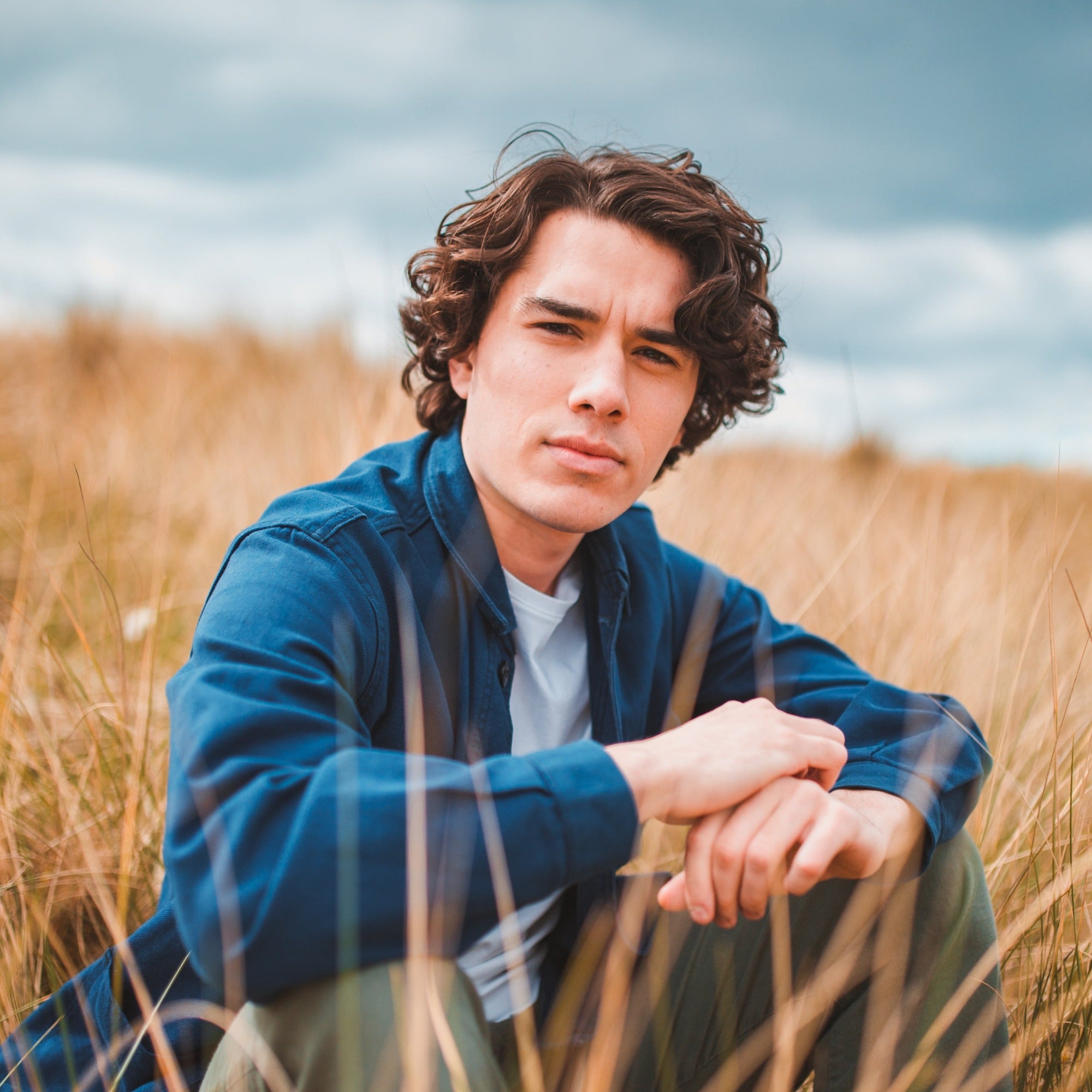 Male model sitting in sand dunes and grass at the beach. He has mid length curly brunette hair and has used Moo and Yoo Sea Salt Spray to style. He is looking directly at the camera and is wearing a blue shirt over a white t shirt.